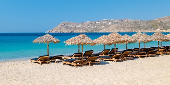 Sandy beach with rows of straw parasols and beach loungers facing a clear blue sea, with a rocky headland in the background under a clear sky.