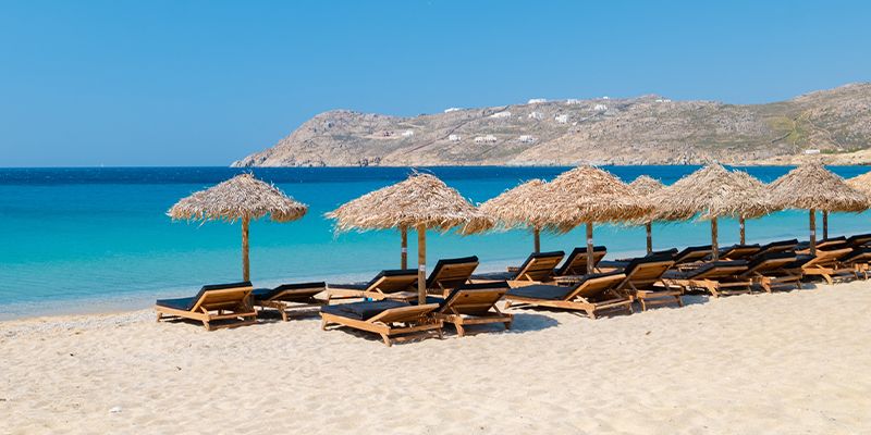 Sandy beach with rows of straw parasols and beach loungers facing a clear blue sea, with a rocky headland in the background under a clear sky.