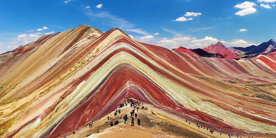 Vibrant striped mountain range with hikers on a trail, set against a blue sky with scattered clouds.
