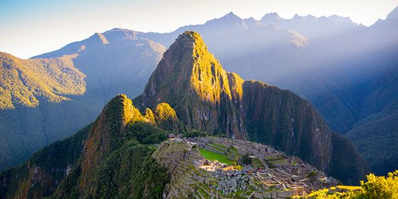 Machu Picchu in sunlight, with terraced ruins and Huayna Picchu mountain in the background, surrounded by lush green Andes mountains.