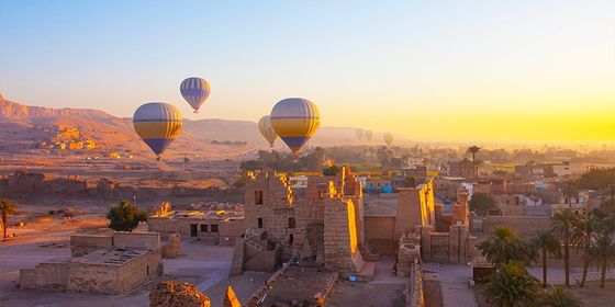 Hot air balloons floating over ancient ruins and desert landscape at sunrise, with distant mountains and a clear sky.