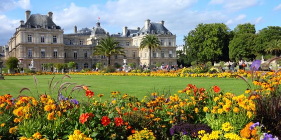 Luxembourg Palace with colorful flower gardens in the foreground, under a blue sky with scattered clouds and visitors walking around.