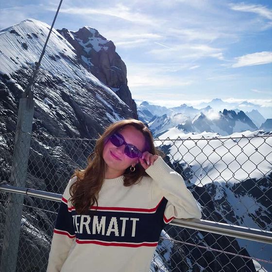 Woman in a "Zermatt" sweater, wearing sunglasses, stands by a railing with a snowy mountain backdrop under a clear blue sky.