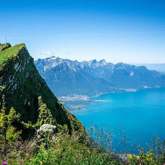 Lush green cliff overlooking a bright blue lake with distant rugged mountains under a clear blue sky.