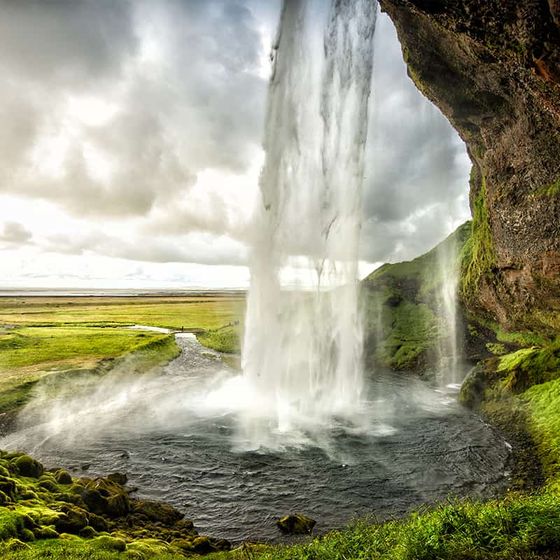 Waterfall cascading over a cliff into a pool, surrounded by lush green landscape and cloudy skies.