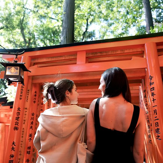 Two women stand in front of red torii gates with Japanese inscriptions, surrounded by lush green trees.