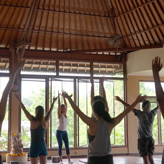 People participating in a yoga class in a spacious room with large windows. An instructor leads with arms raised; lush greenery is visible outside.