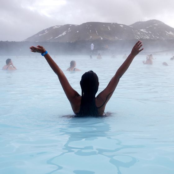 Person with raised arms enjoying a geothermal pool, surrounded by mist, with mountains in the background.