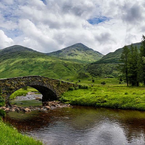 Stone bridge over a tranquil river, surrounded by lush green hills and trees under a partly cloudy sky.