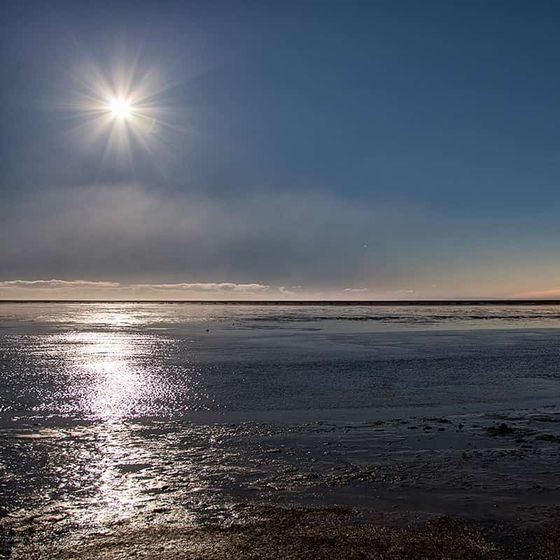 Sunset over a tranquil beach with a bright sun and clear blue sky reflecting on wet sand and calm water.