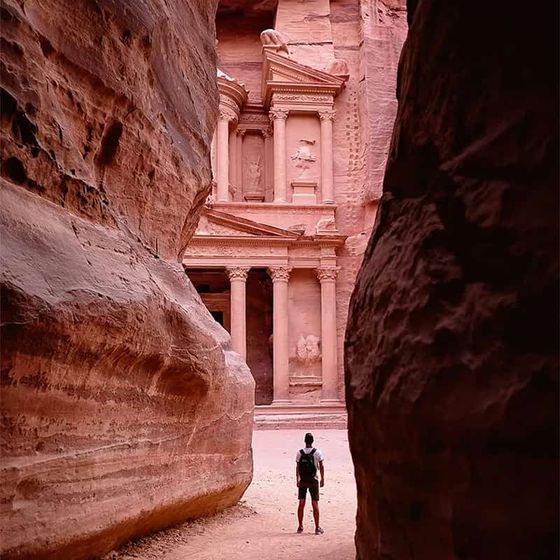 A person with a backpack stands before the ancient, carved rock facade of Al-Khazneh in Petra, Jordan, framed by towering canyon walls.