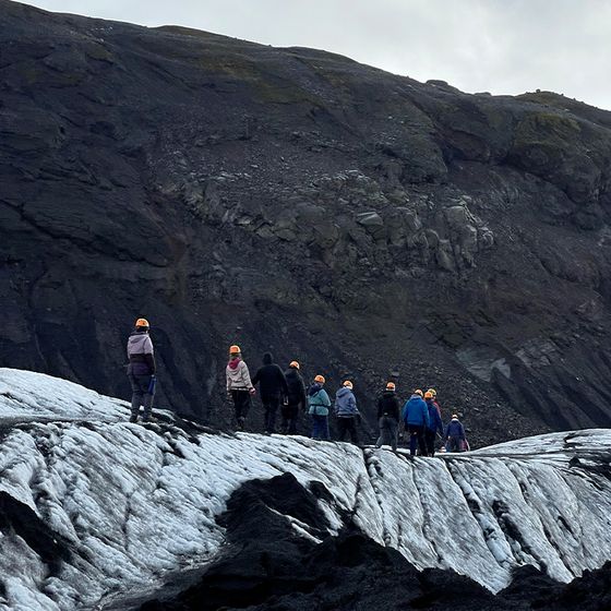 A group of people wearing helmets hike across a glacier surrounded by rocky terrain under a cloudy sky.