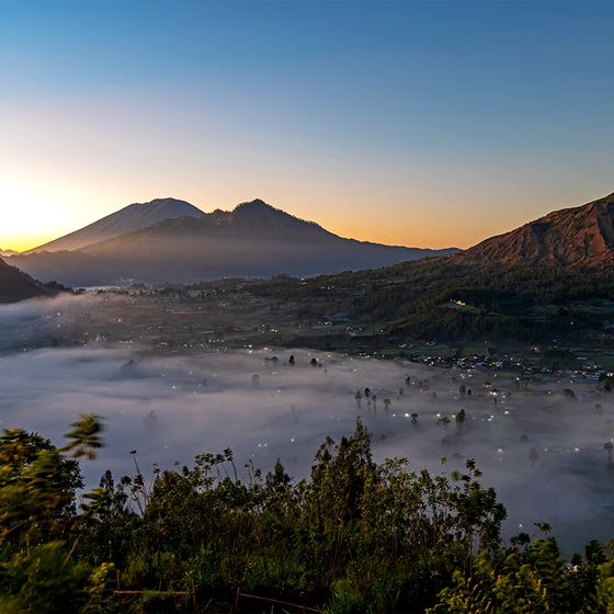 Scenic sunrise over misty valley with Mount Batur in the background, surrounded by lush green hills and a faintly visible village below.