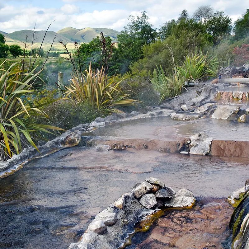Steaming natural hot springs with rock formations, surrounded by lush greenery and distant rolling hills under a partly cloudy sky.