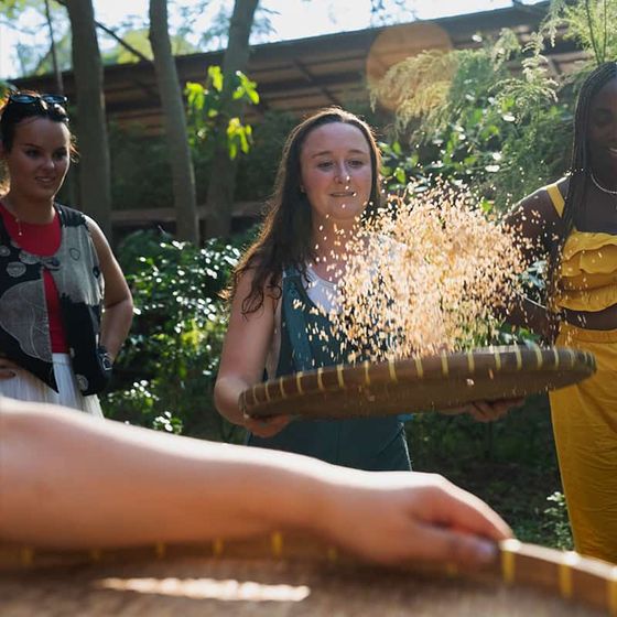 Four people outdoors, one using a bamboo tray to sift grains. Sunlight filters through trees, creating a serene setting.