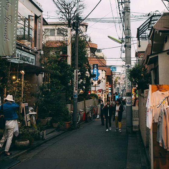 A narrow urban street lined with shops and plants, with people walking and electrical wires overhead on a cloudy day.