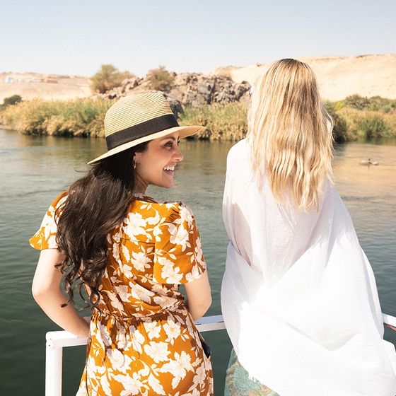 Two women on a boat enjoying a sunny day on a river, with one wearing a straw hat and floral dress, overlooking lush greenery and rocky hills.