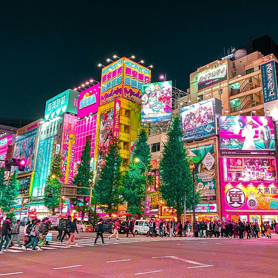 Vibrant city street at night, featuring colorful neon-lit buildings, busy crosswalk, and groups of pedestrians under a dark sky.