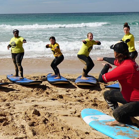 Four people in yellow shirts practice surfing techniques on boards in the sand, guided by an instructor in red, near ocean waves.