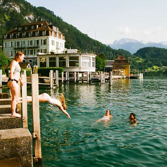 People swimming and diving into a clear lake near buildings, surrounded by mountains on a sunny day.