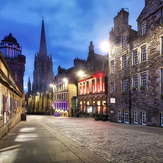 Cobblestone street in Edinburgh at dusk, lined with historic buildings, warm lights, and a tall spire in the background.