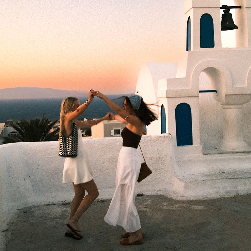 Two women joyfully dance on a rooftop at sunset, with a white dome and the sea in the background.