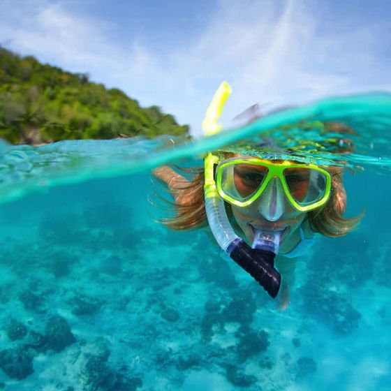 Person snorkeling in clear turquoise water near a lush green island, wearing a yellow mask and snorkel, with vibrant coral visible below.