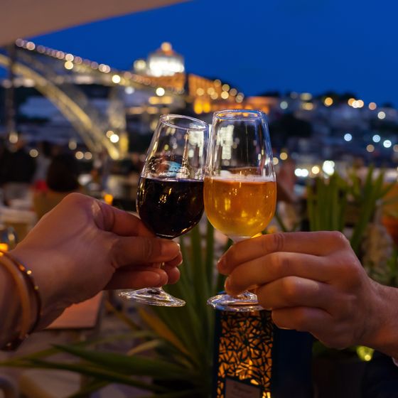 Two hands clinking glasses of red and white wine against a backdrop of a lit-up bridge and cityscape at night.