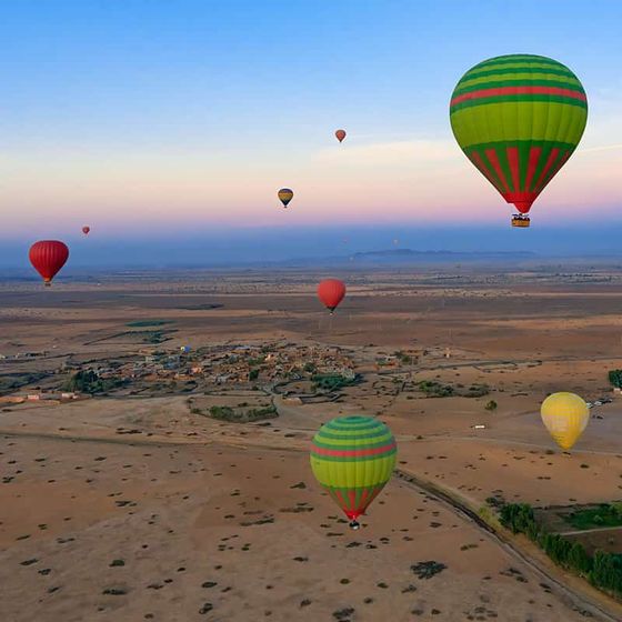 Colorful hot air balloons floating over a vast desert landscape at sunrise, with scattered villages and mountains in the distance.