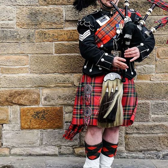 A person in traditional Scottish attire plays bagpipes in front of a stone wall. The outfit features a tartan kilt and ornate details.