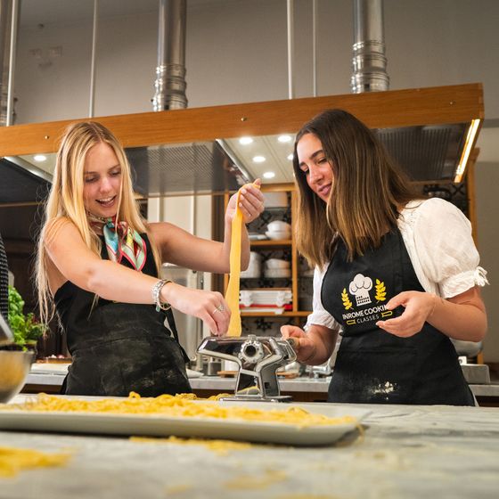 Two women making pasta using a pasta machine in a modern kitchen. Both are smiling and wearing aprons, with fresh pasta on the counter.