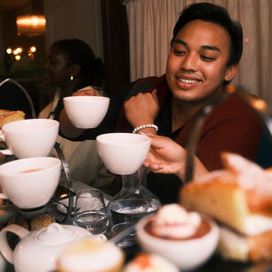 A person smiles while reaching for a teacup in a cozy setting with various pastries and another person in the background.