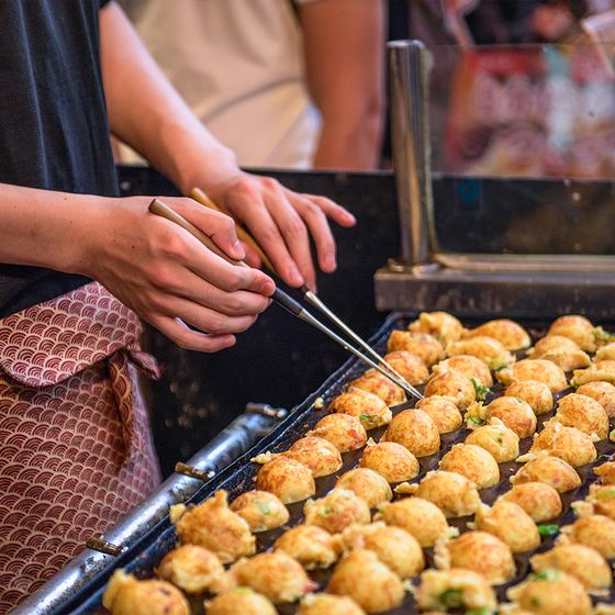 Close-up of a person cooking takoyaki on a hot grill, using chopsticks to turn the round, golden-brown balls in an outdoor setting.
