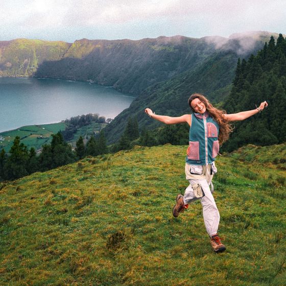 A joyful person with long hair jumps on a grassy hillside, with a scenic view of a lake and green mountains in the background.