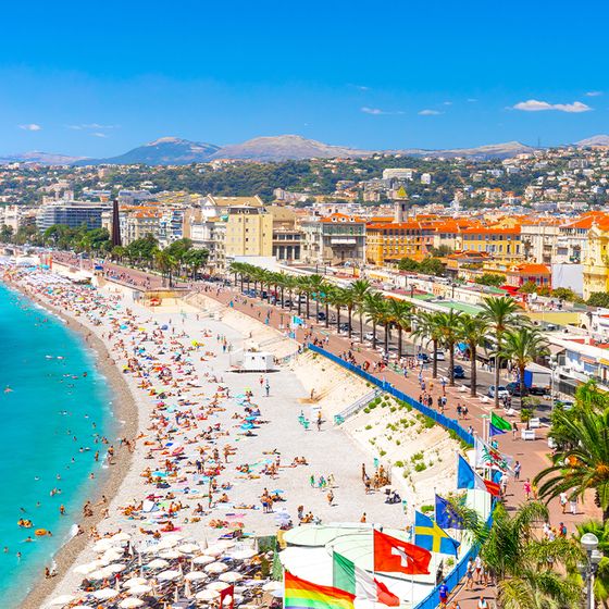 A vibrant beach in Nice, France, with turquoise water, people sunbathing and swimming, palm trees, flags, and colorful buildings under a clear blue sky.
