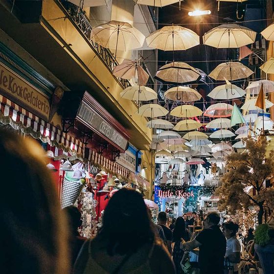 A bustling night market scene with crowds under a ceiling of hanging umbrellas and vibrant shop displays.