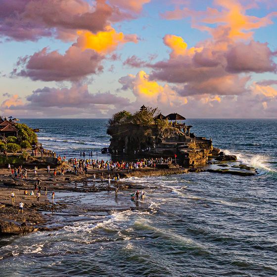 Crowd gathered on rocky shore near Tanah Lot Temple in Bali at sunset, with vibrant clouds and ocean waves creating a picturesque scene.