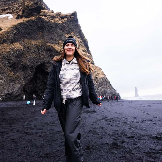 A person smiles standing on a black sand beach with rugged cliffs in the background under a cloudy sky.