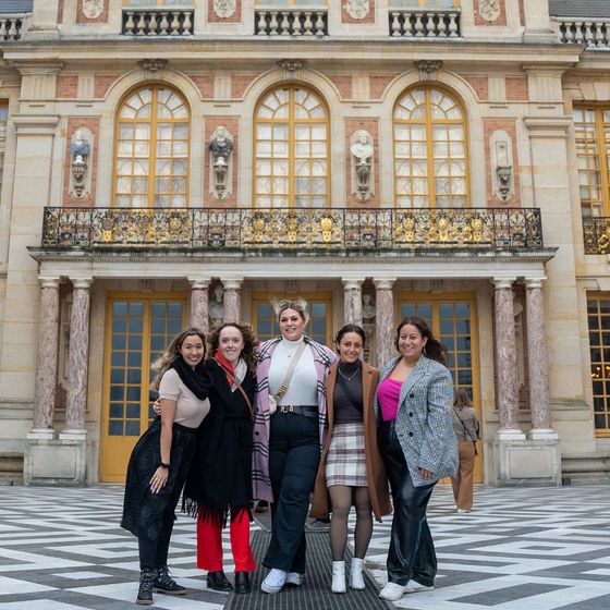 Five people smiling in front of an ornate building with large windows, stone columns, and checkered floor.
