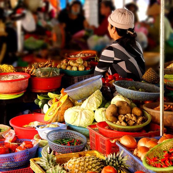 A vendor sits at a vibrant market stall selling vegetables and fruits, including cabbages, potatoes, and pineapples, in baskets.