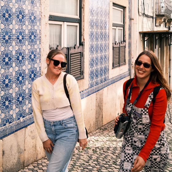 Two women smiling in sunglasses walk on a cobblestone street with blue patterned tiles on the wall in a sunny setting.