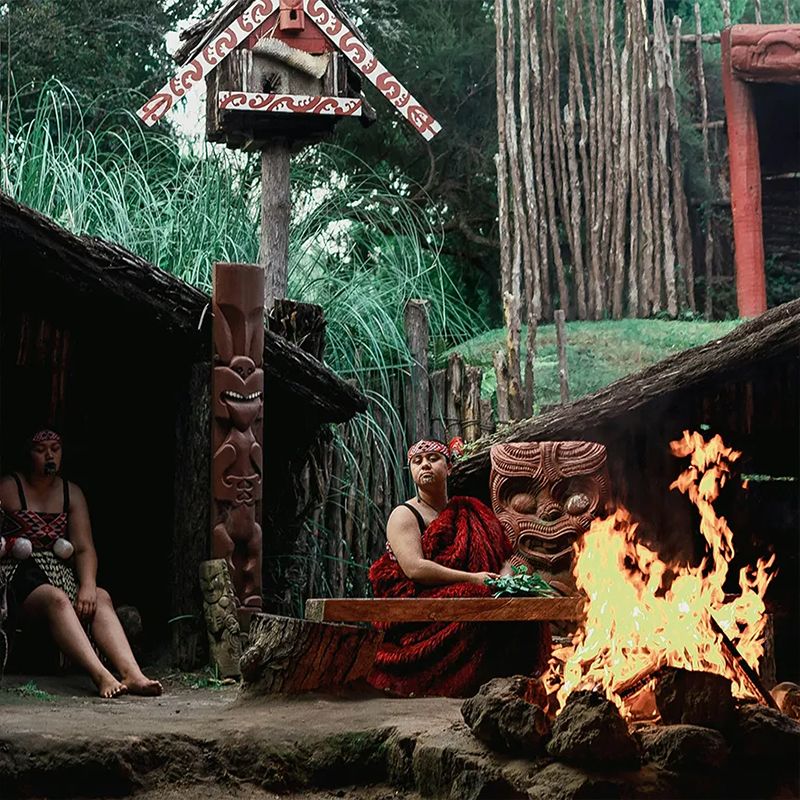 Two people in traditional attire by a large fire, surrounded by carved wooden structures and lush greenery in a cultural setting.