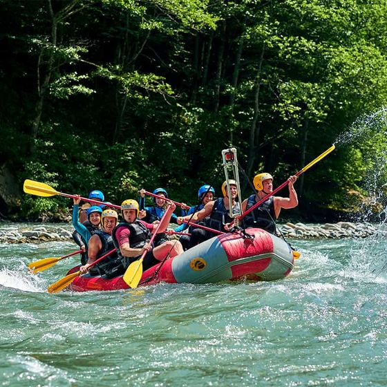 A group of people wearing helmets and life vests are white-water rafting on a river, paddling through rapids surrounded by lush greenery.