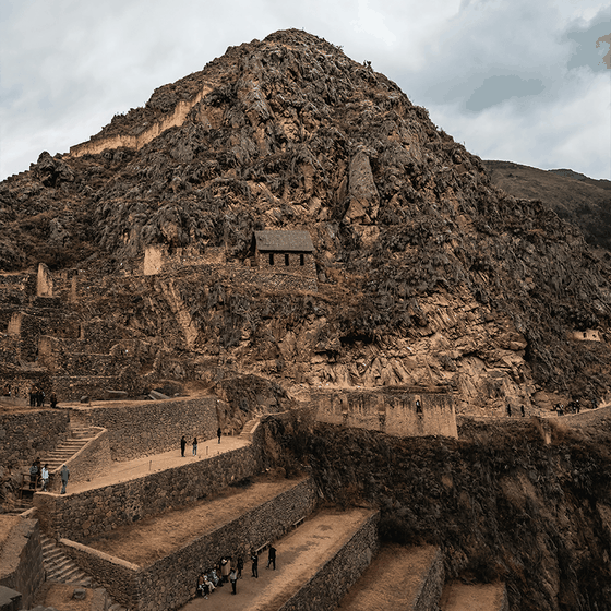 Ancient stone terraces and a small building nestled against a rugged mountain slope, with scattered people exploring the site.