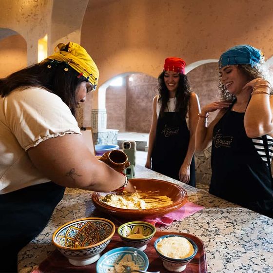 Three women in colorful headscarves and aprons joyfully prepare food together at a countertop with bowls and ingredients.
