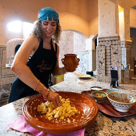 Woman kneading dough in a rustic kitchen, wearing a blue headscarf and black apron, surrounded by colorful bowls and decorative tiles.
