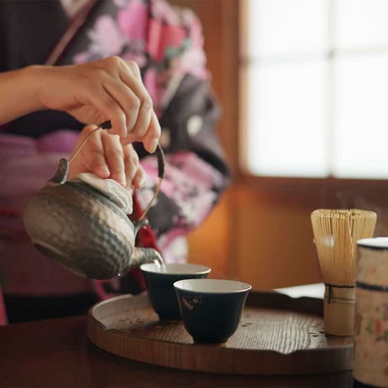 A person in a kimono pours tea from a metal teapot into small cups on a wooden tray, with a bamboo whisk and a patterned container nearby.