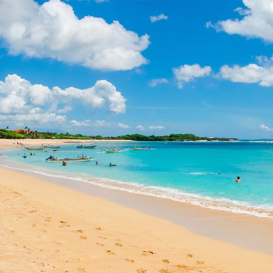 A sunny beach scene with turquoise waters, several boats, people swimming, and footprints in the sand under a blue sky with fluffy clouds.