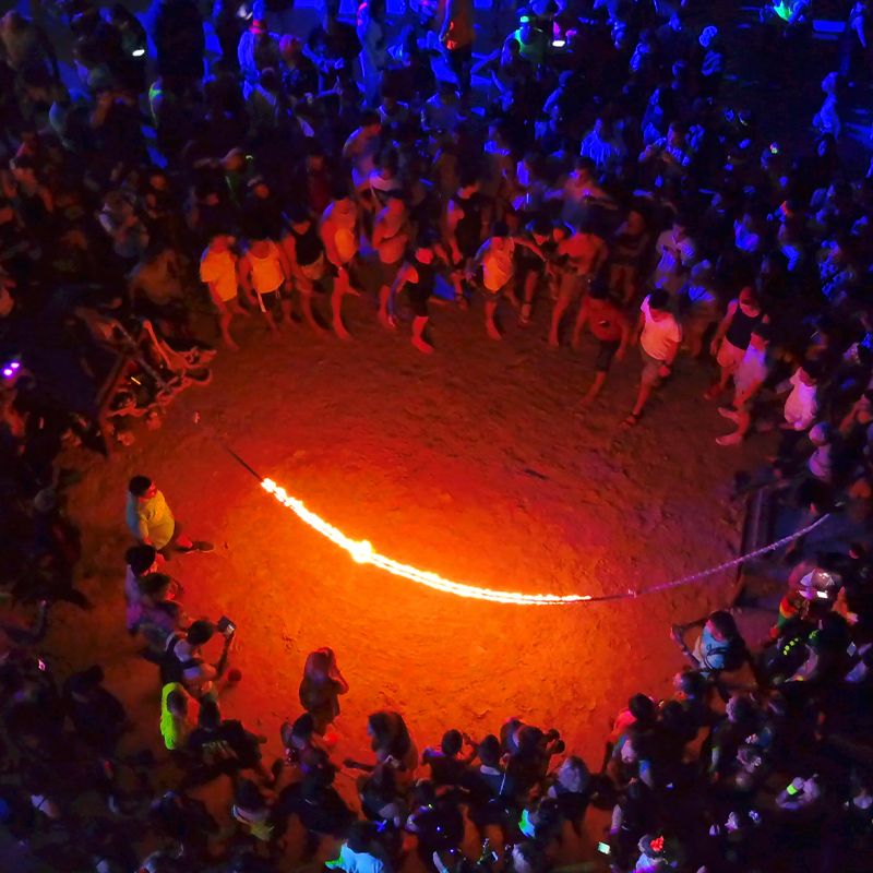 Aerial view of a lively night gathering with people surrounding a flaming jump rope on sandy ground, illuminated by vibrant orange and blue lights.