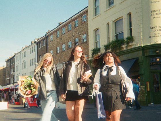 Three friends walk down a sunny street; one holds flowers. They're smiling, with shops and a brick building in the background.
